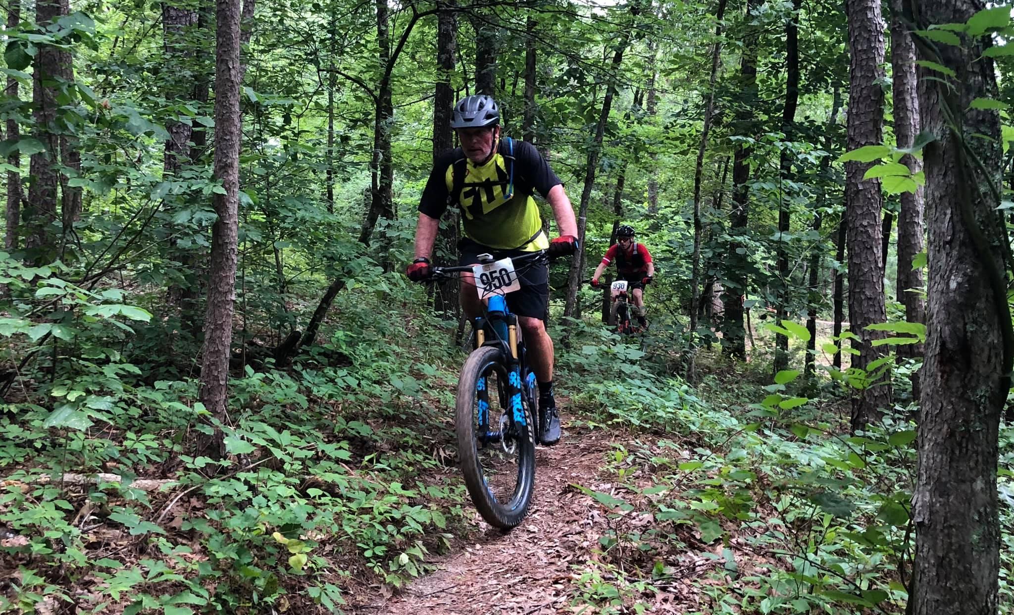 A mountain biker wearing a yellow and black jersey navigates a narrow dirt trail through a dense forest, with lush greenery surrounding the path. Another biker in a red jersey follows closely behind. Barber Hills Trail at Pat Mayse Lake mountain bike trail.