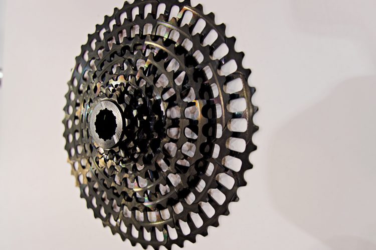 Close-up image of a bicycle cassette, showcasing its intricate design with multiple gears arranged in a circular pattern. The cassette has metallic black and iridescent elements, highlighting the teeth and central hub. The background is plain, emphasizing the details of the cassette.
