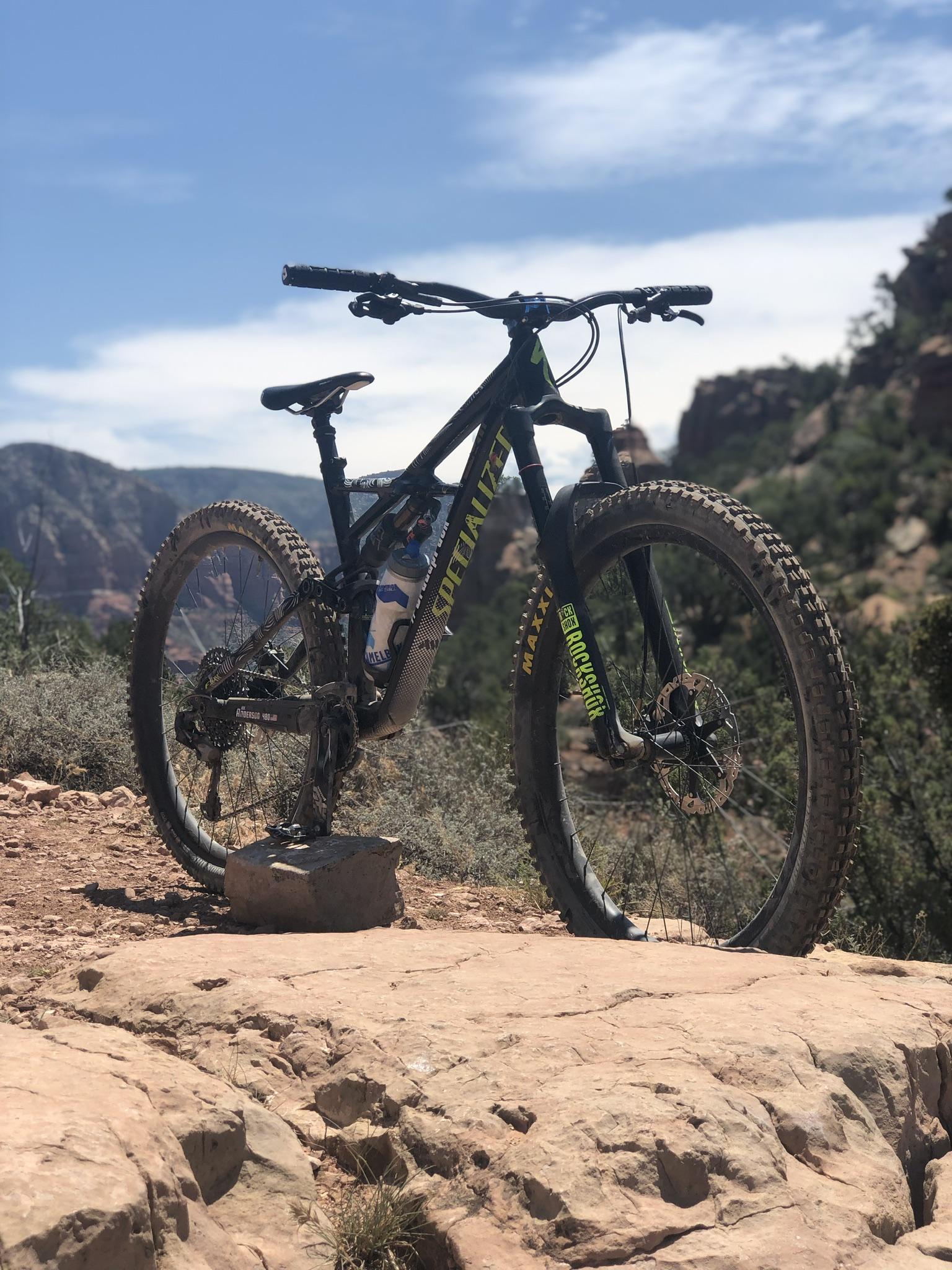 Specialized Enduro SL Comp FSR: A black mountain bike with bright yellow accents is positioned on a rocky terrain, surrounded by greenery and mountains in the background. The bike has a water bottle attached and is resting on a large stone, with a clear blue sky above.