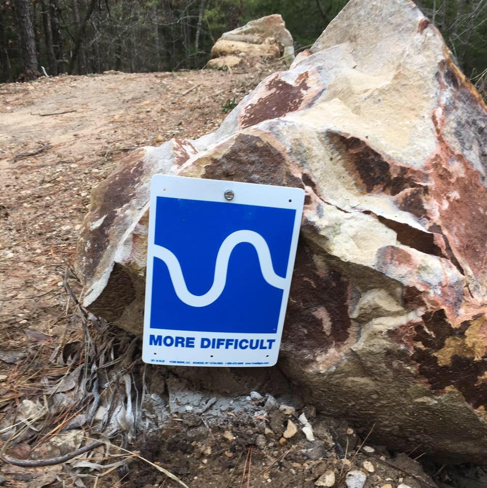 A blue and white trail marker sign that reads "MORE DIFFICULT," placed next to a large rock in a wooded area. The background features a dirt path and trees. Barber Hills Trail at Pat Mayse Lake mountain bike trail.
