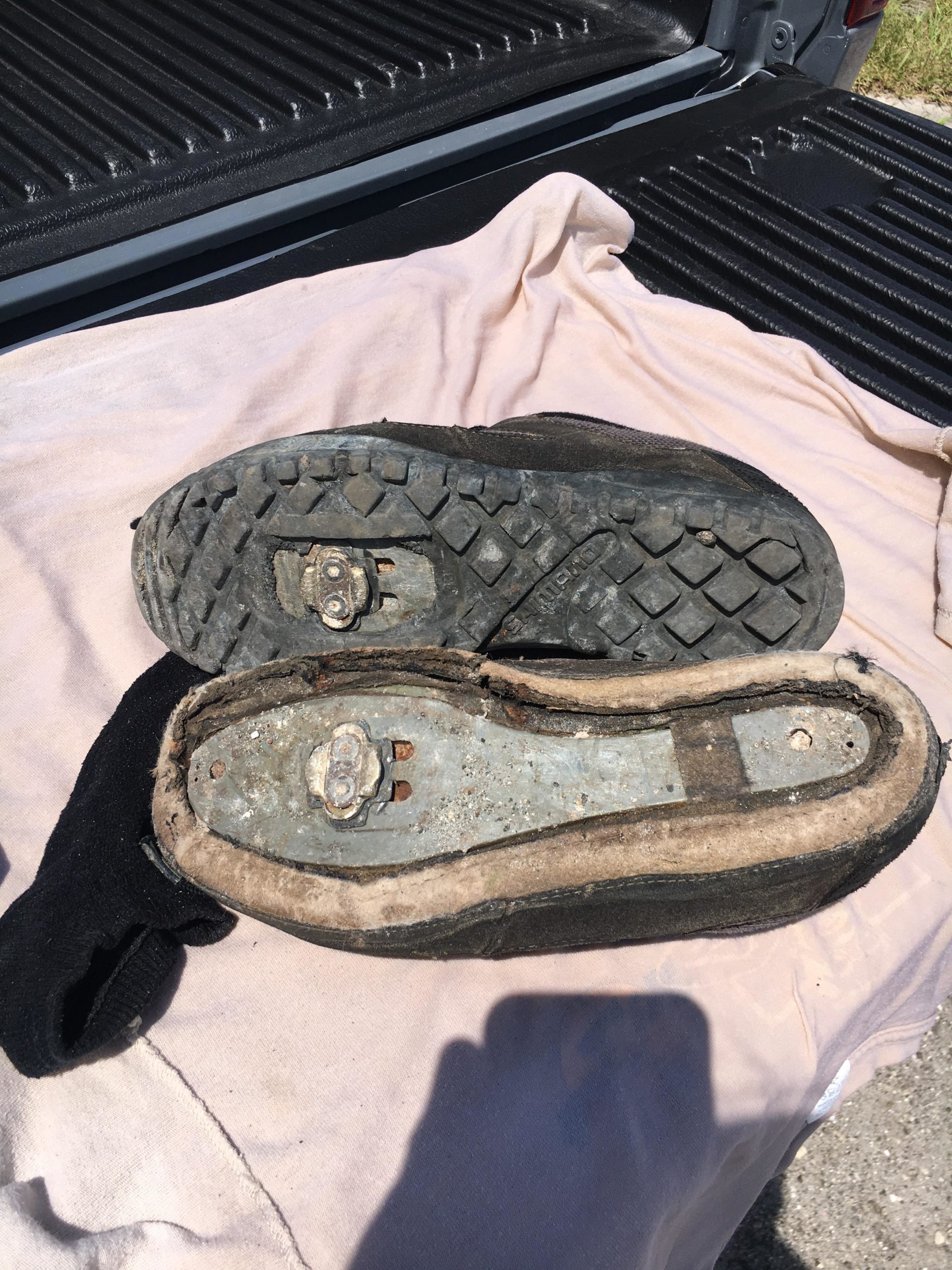 Close-up image of a pair of worn-out shoes with visible wear and tear, displayed on a light cloth. The soles show signs of use, including dirt and scuff marks, while the inner part of one shoe is partially exposed. The background is blurred, featuring the texture of a truck bed. Balm Boyette Scrub Preserve mountain bike trail.