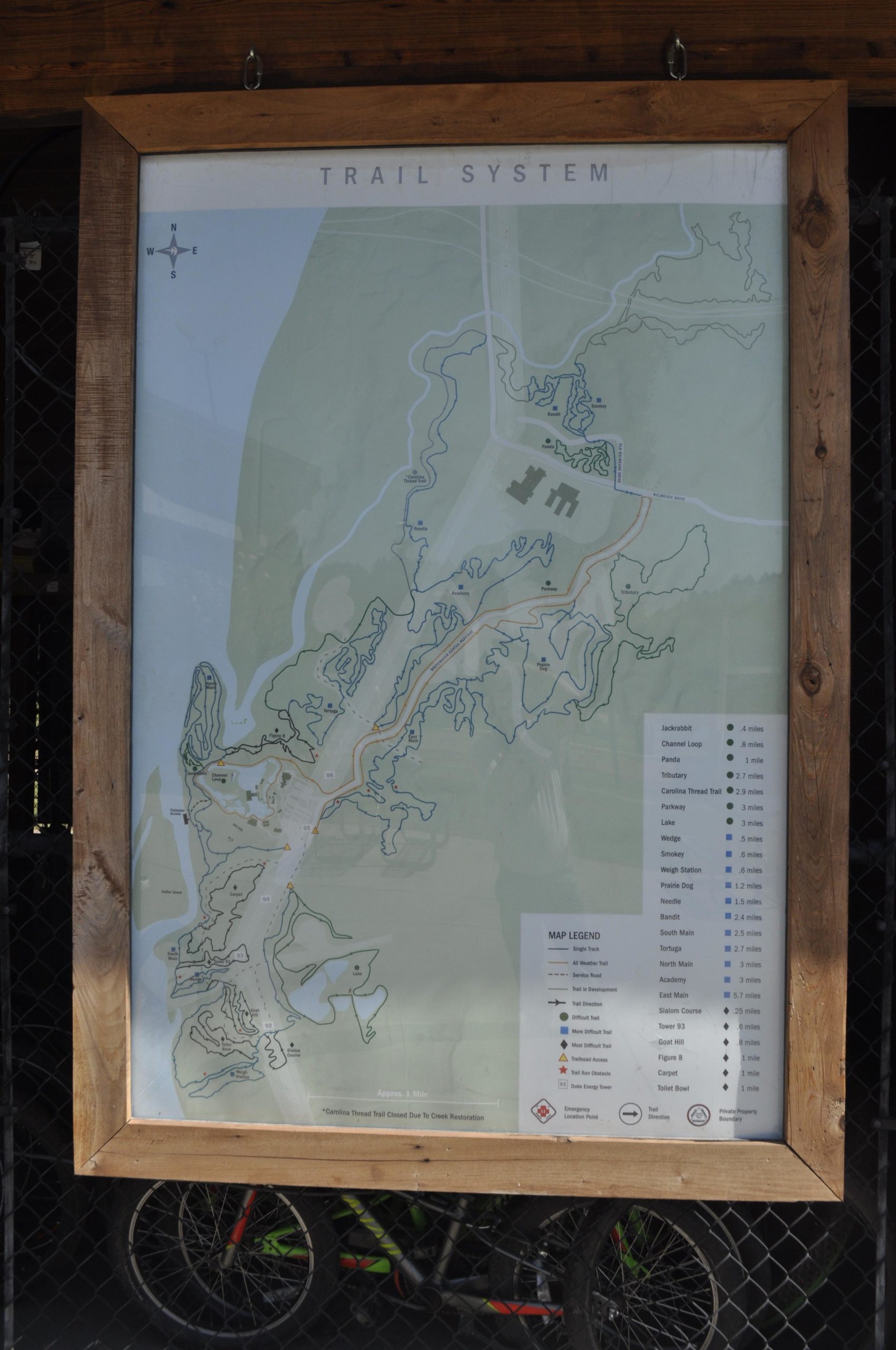 Map of a trail system, featuring various marked trails, distances, and a legend for navigation. The map includes details such as specific trail names, difficulty levels, and nearby points of interest, all framed in rustic wood. In the background, bicycles are partially visible. USNWC mountain bike trail.