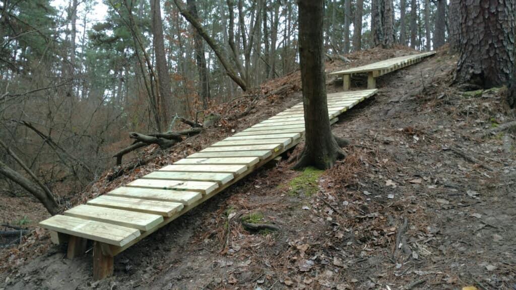 A wooden pathway traversing a sloped, natural area in a forest, surrounded by trees and underbrush. The boardwalk is constructed of wide wooden planks, providing a clear route through the uneven terrain. Barber Hills Trail at Pat Mayse Lake mountain bike trail.