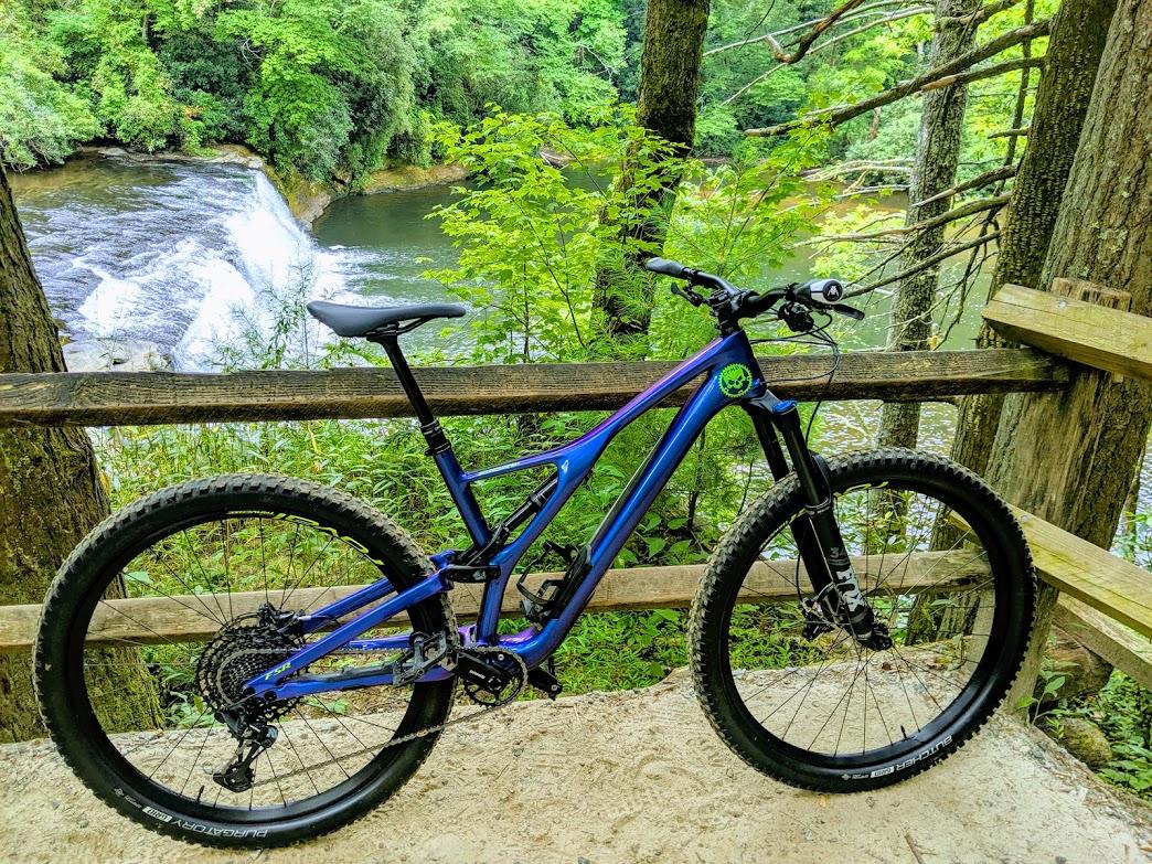 Specialized Stumpjumper FSR Comp Carbon 29: Mountain bike resting on a wooden railing, with a backdrop of a river and waterfall surrounded by lush greenery.