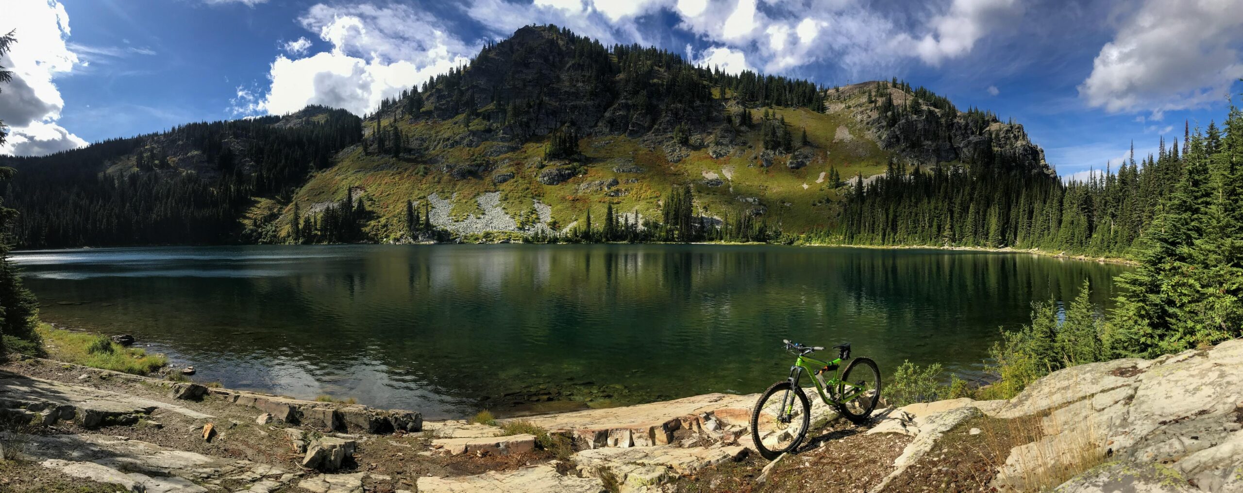 Giant Trance: A scenic view of a tranquil lake surrounded by lush green hills and dense pine forests. In the foreground, a green mountain bike is parked on a rocky shoreline, reflecting the vibrant colors of the landscape. The sky is bright with fluffy clouds, and the water’s surface shimmers under sunlight, creating a peaceful and inviting outdoor atmosphere.