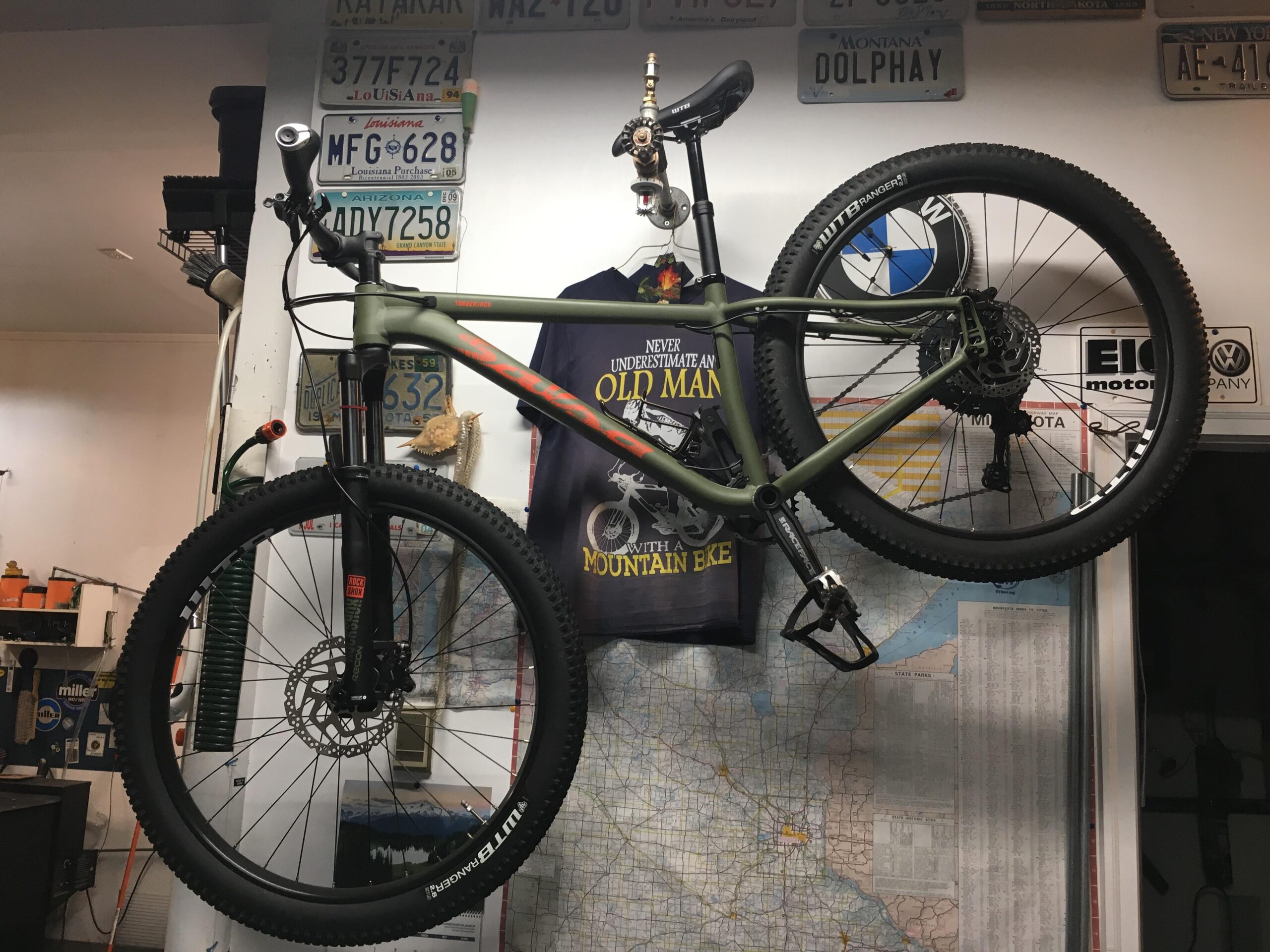 Salsa Timberjack: A mountain bike hanging on a wall in a garage or workshop, surrounded by various vintage license plates and a map. A T-shirt with a graphic design featuring a bike is displayed nearby, along with some tools and bicycle accessories. The bike has a green frame and rugged tires, indicating it is designed for off-road use.