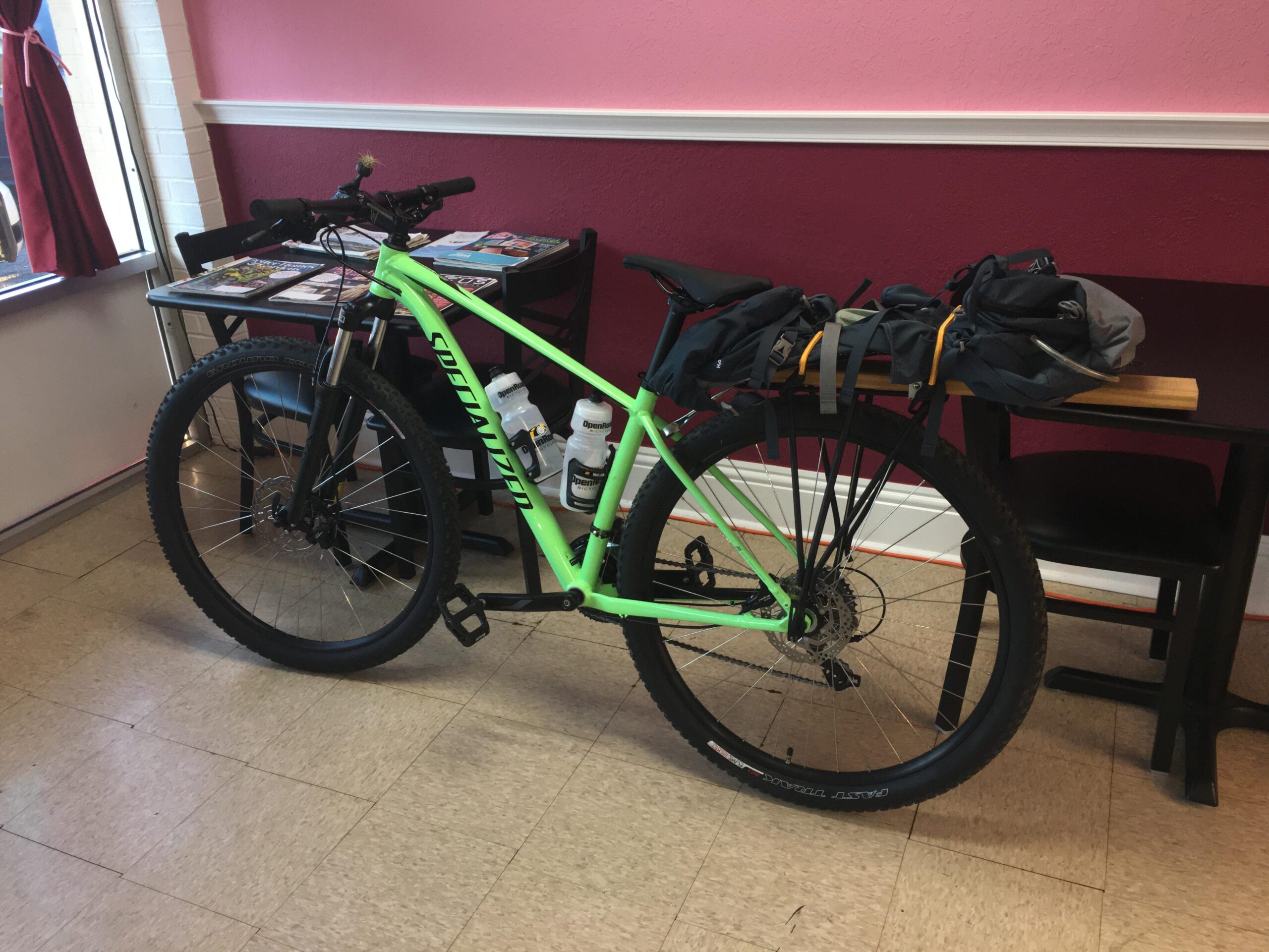 Specialized Rockhopper 29: A bright green mountain bike is leaning against a small black table inside a room with pink walls. The bike has a backpack and some gear attached to it, and there are water bottles on the floor nearby. A window with red curtains can be seen in the background.
