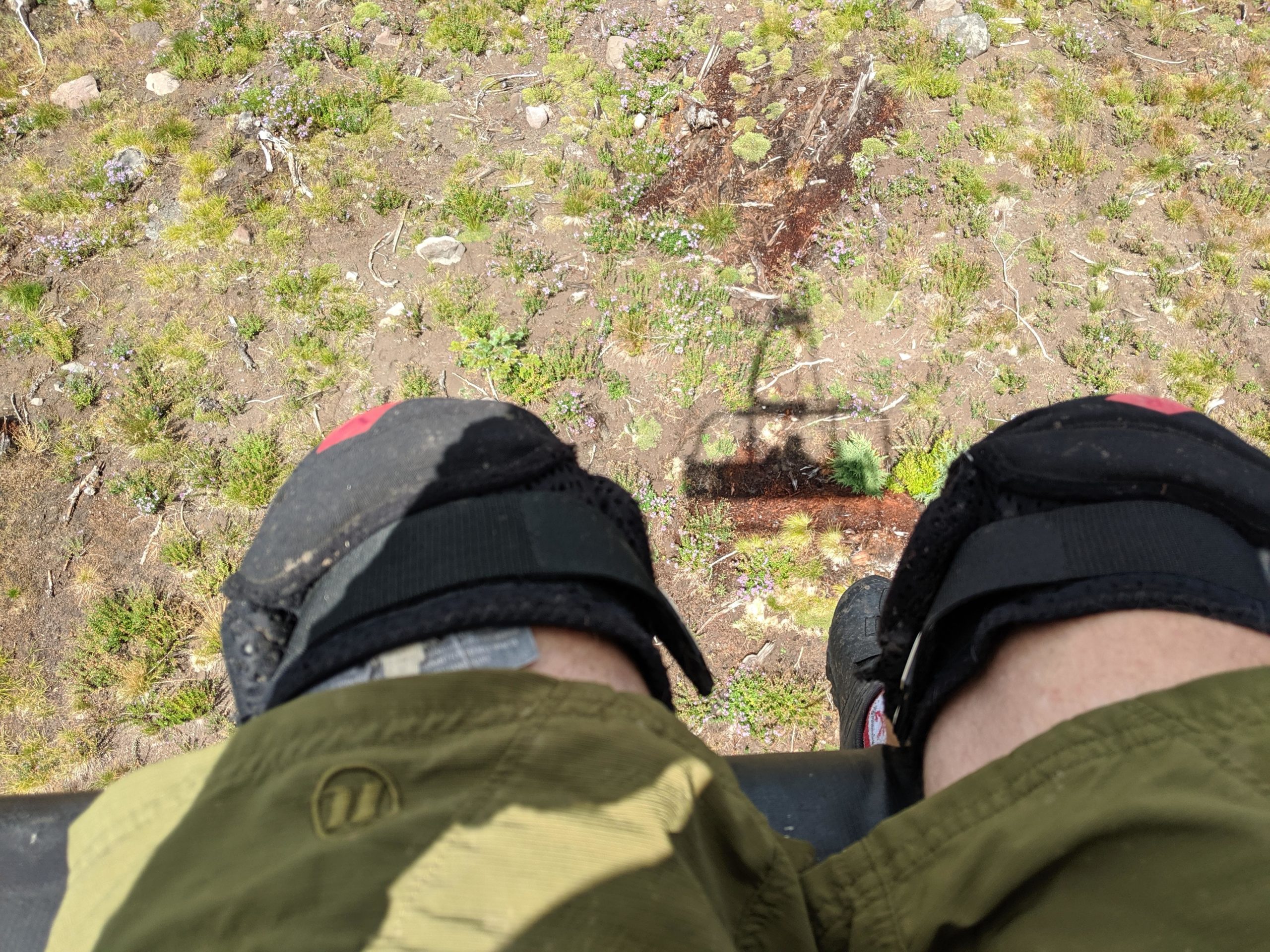 A close-up view of two legs wearing knee pads and shorts, suspended above a grassy landscape. The ground below features patches of dirt, small plants, and rocks, while a shadow from an overhead cable is visible. Timberline Bike Park mountain bike trail.