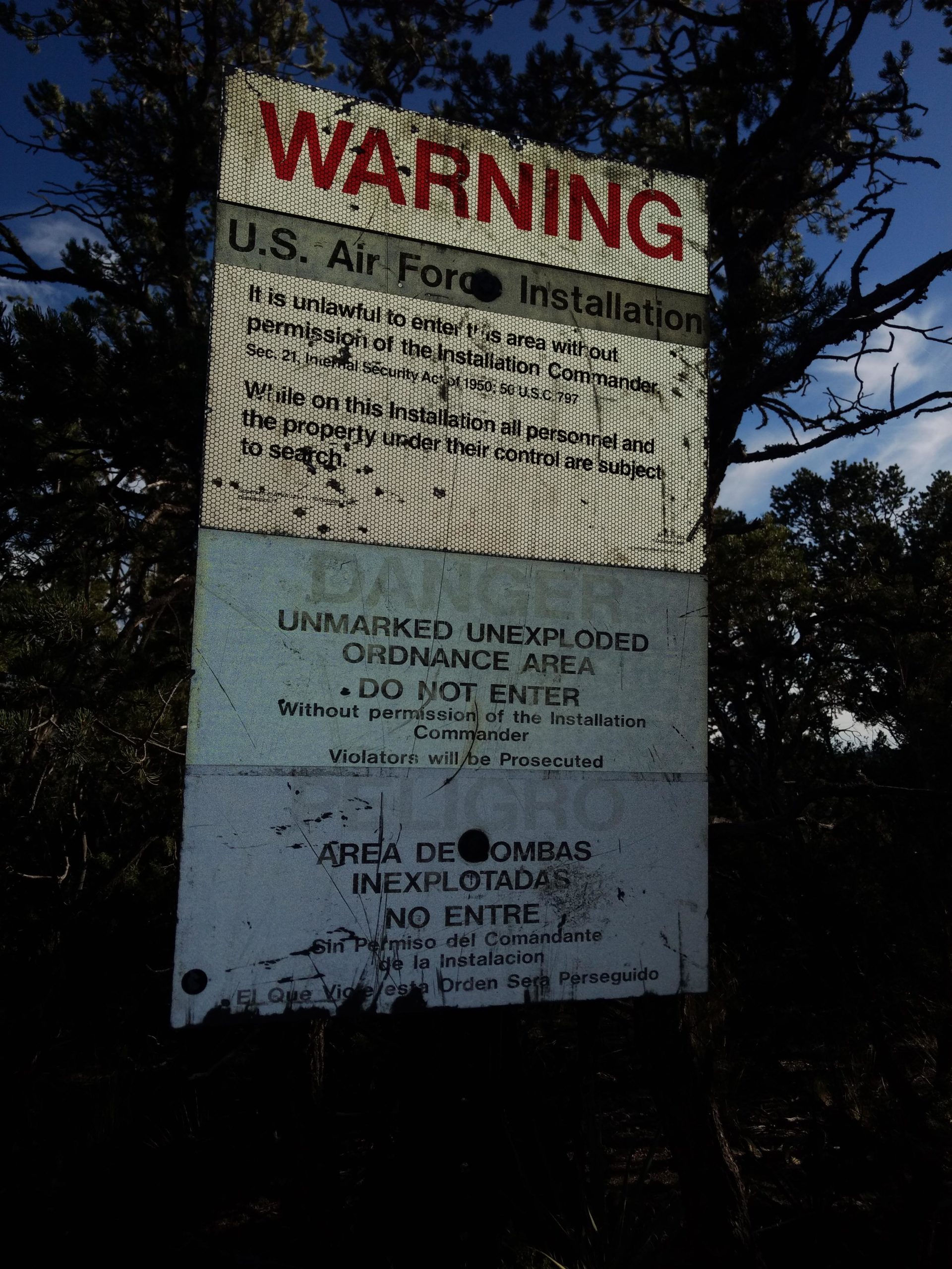 A weathered warning sign indicating a U.S. Air Force installation. The sign states that entry is prohibited without permission, highlights the presence of an unmarked unexploded ordnance area, and notes that violators will be prosecuted. The text is presented in English and Spanish, with cautionary language regarding safety. The background features trees and natural vegetation. Tunnel Canyon mountain bike trail.