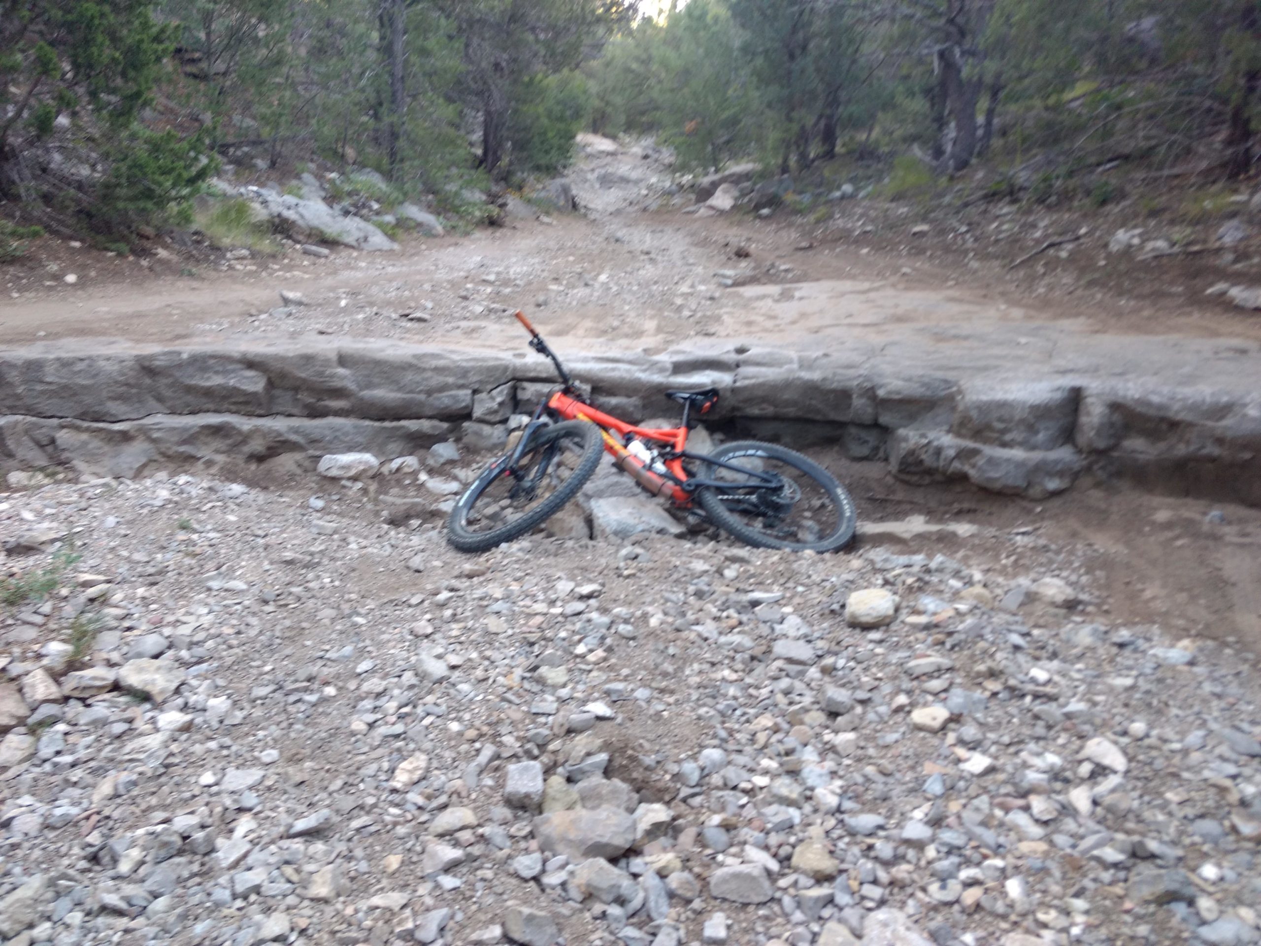 A mountain bike lying on its side on a rocky trail, next to a stone structure. The surrounding area is filled with dirt and small rocks, and trees are visible in the background. Otero Canyon mountain bike trail.