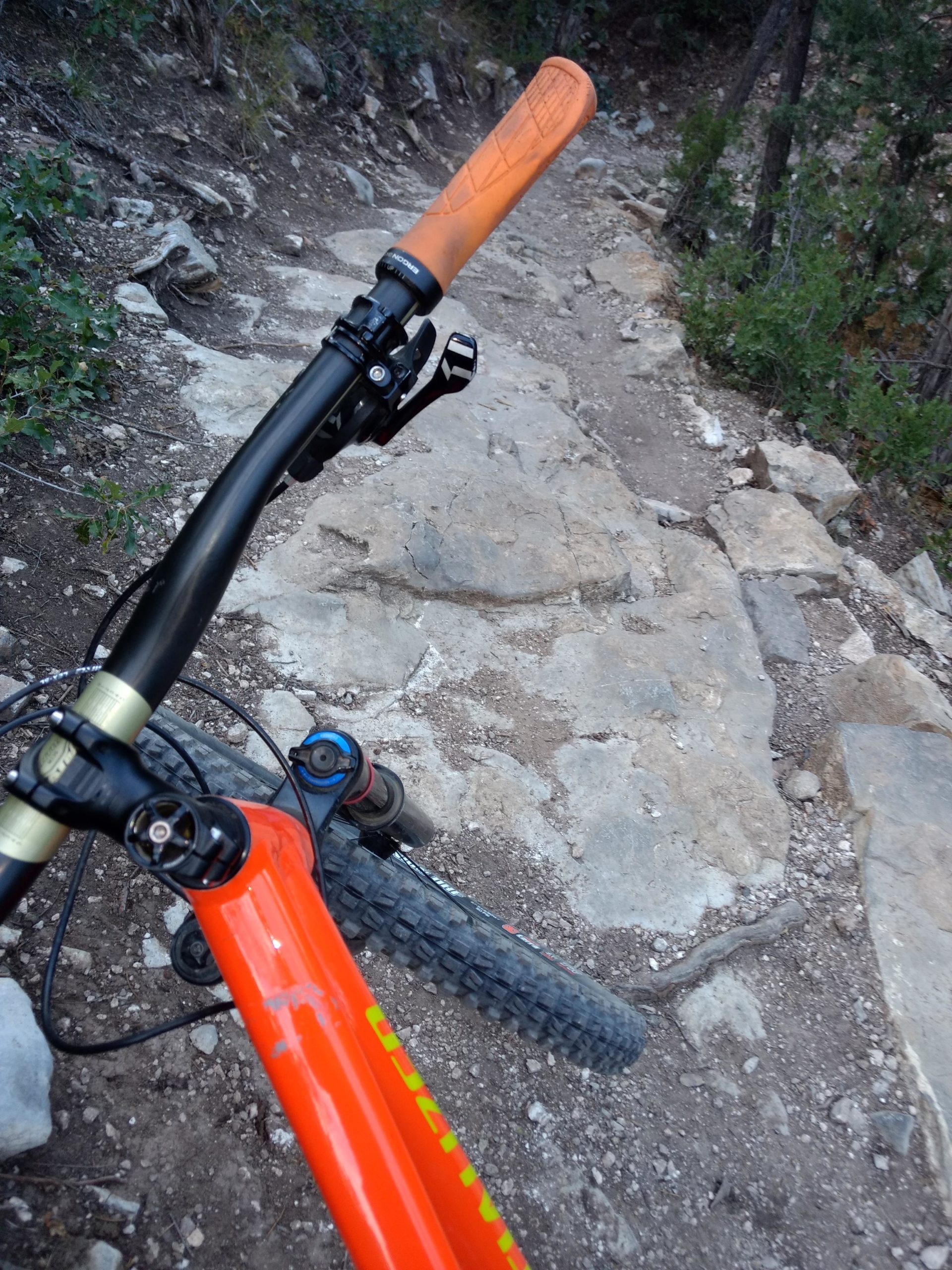 Close-up view of a mountain bike handlebar with an orange grip, positioned above a rocky dirt trail surrounded by trees. The bike frame is bright orange, and the scene suggests an outdoor cycling adventure in a natural setting. Tunnel Canyon mountain bike trail.