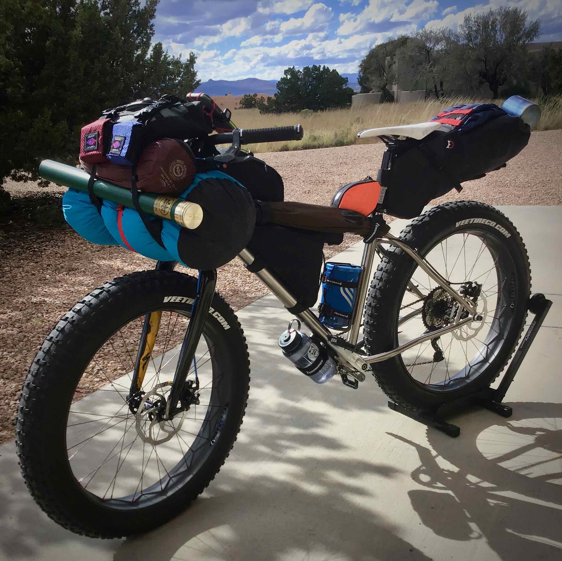 Motobecane Motobecane Bullet Titanium Fat bike: A fat bike equipped for bikepacking, featuring various bags and gear secured on the frame. The bike is parked on a concrete surface with a natural landscape in the background, including trees and distant mountains under a partly cloudy sky.