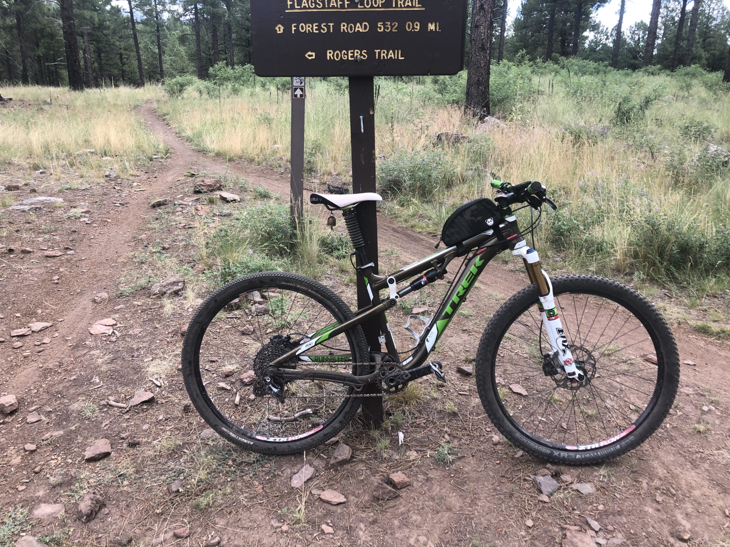 A mountain bike parked next to a trail sign in a wooded area. The sign marks the Flagstaff Loop Trail and nearby routes, with a mix of dirt and rocky terrain visible. Lush green grass and trees surround the scene, indicating a natural outdoor environment. Soldiers Loop / Fairgrounds trails mountain bike trail.