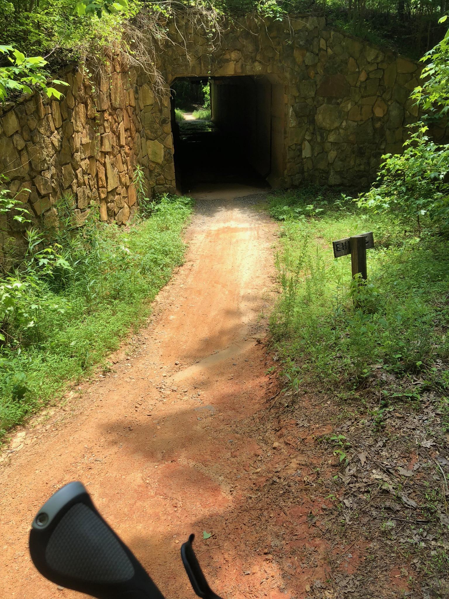 A dirt path leading towards a stone tunnel surrounded by lush greenery. A wooden sign labeled "End" is visible on the right side of the pathway. The scene is illuminated by natural sunlight filtering through the trees. Anne Springs Close Greenway mountain bike trail.