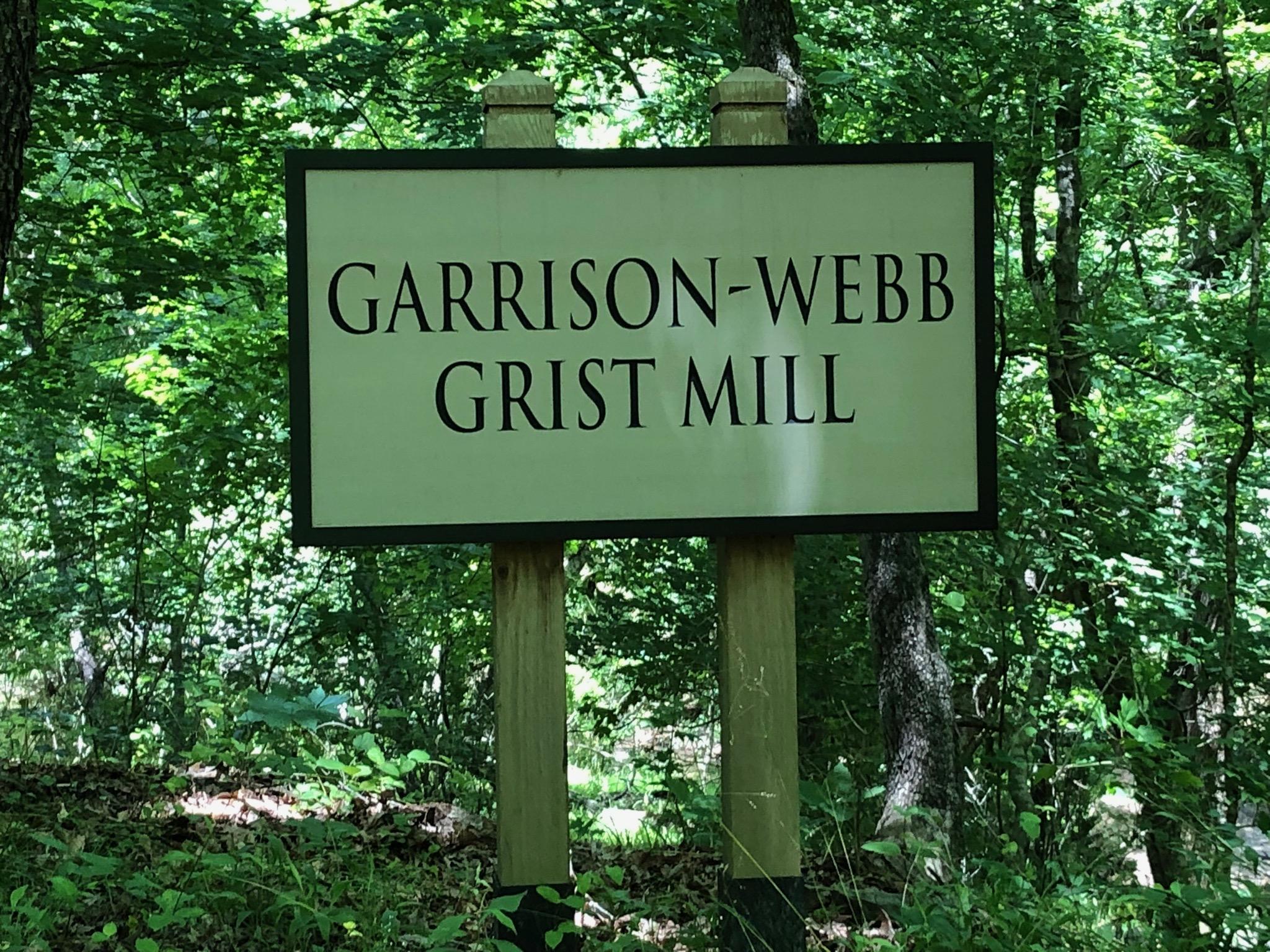 A wooden sign that reads "Garrison-Webb Grist Mill," surrounded by dense green foliage and trees in a natural setting. Anne Springs Close Greenway mountain bike trail.