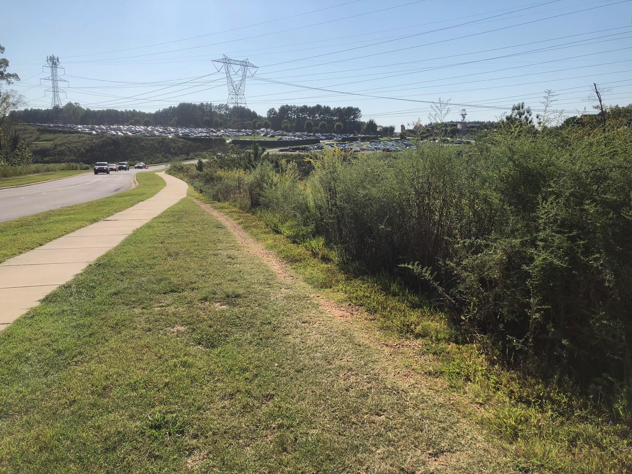 A view of a paved sidewalk winding alongside a road, bordered by lush greenery and bushes. In the background, several power lines stretch across the sky, while a line of parked cars is visible on a hillside. The scene is set in bright daylight with clear blue skies. USNWC mountain bike trail.