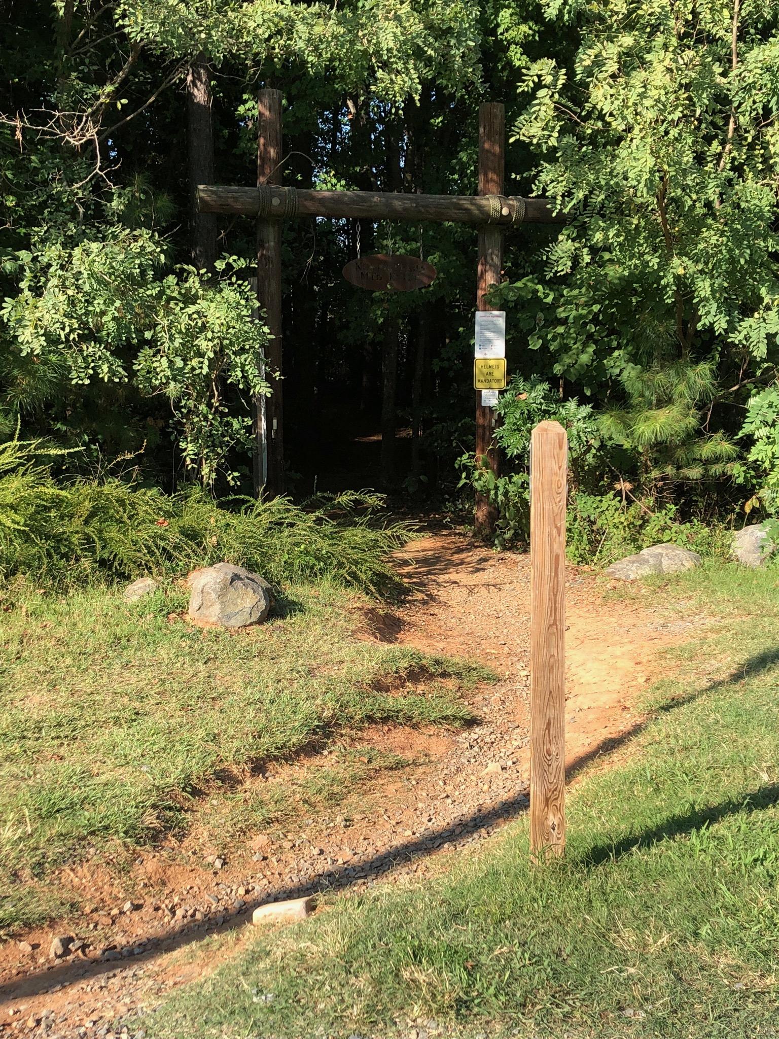 A wooden entrance sign stands at the edge of a dirt path leading into a wooded area. Lush greenery surrounds the entrance, with a small post indicating the trail. Sunlight filters through the trees, creating a serene and inviting atmosphere. USNWC mountain bike trail.