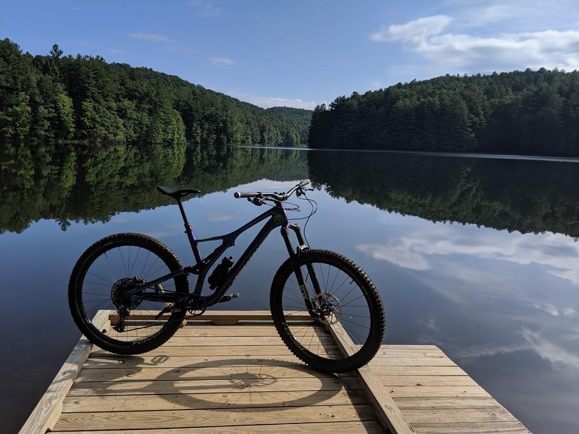 A mountain bike is parked on a wooden dock by a serene lake, surrounded by dense green trees and reflecting perfectly in the calm water. The sky is clear with a few clouds, creating a peaceful outdoor scene. DuPont State Forest mountain bike trail.