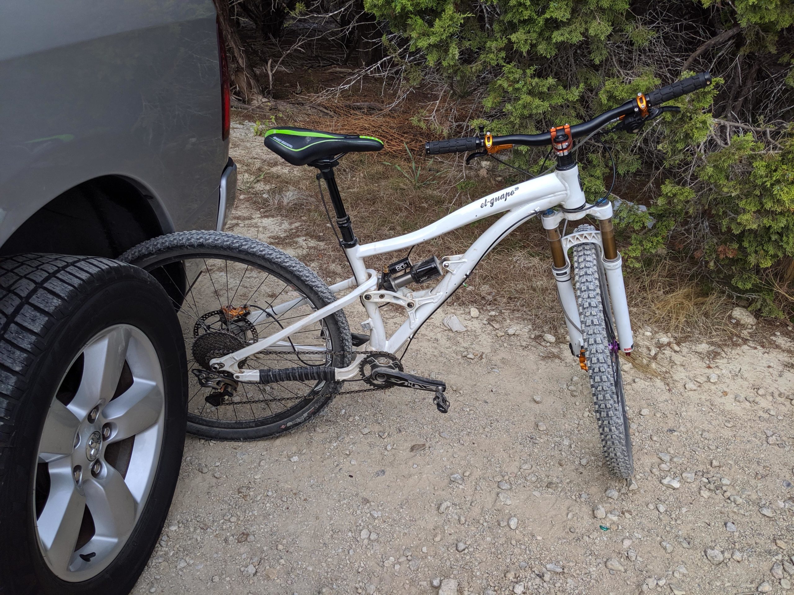 A white mountain bike leaning against the side of a gray vehicle, partially obscured by the tire. The background features a dirt pathway with scattered rocks and greenery. Dana Peak mountain bike trail.