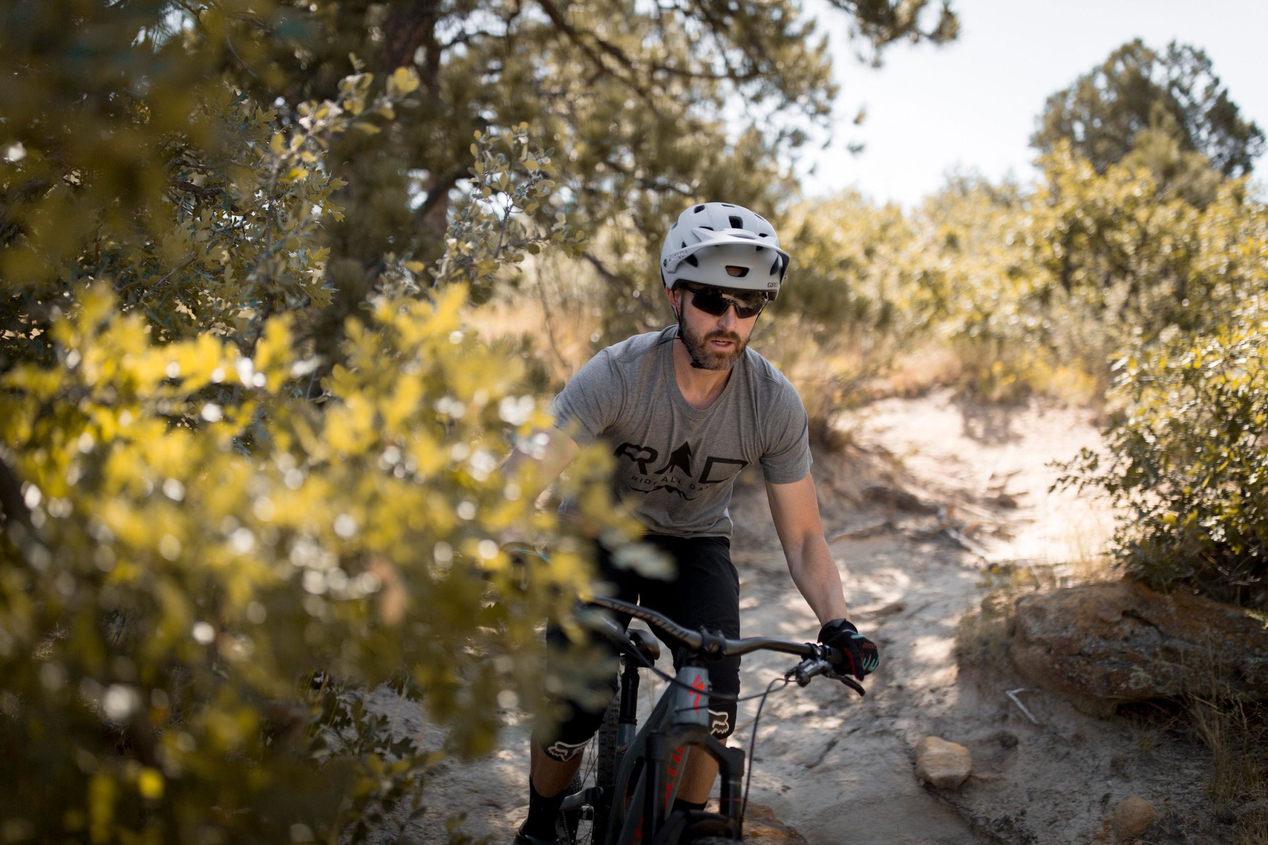 A mountain biker wearing a helmet and sunglasses rides along a rocky trail surrounded by greenery, focusing on navigating the terrain. Palmer Park mountain bike trail.