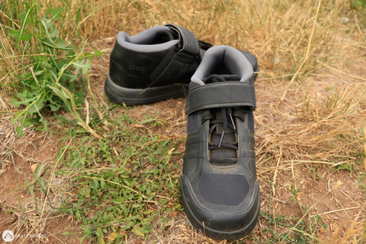 A pair of black hiking shoes resting on the ground among grass and dried foliage. The shoes feature a sturdy design with a Velcro strap and mesh material, appropriate for outdoor activities.