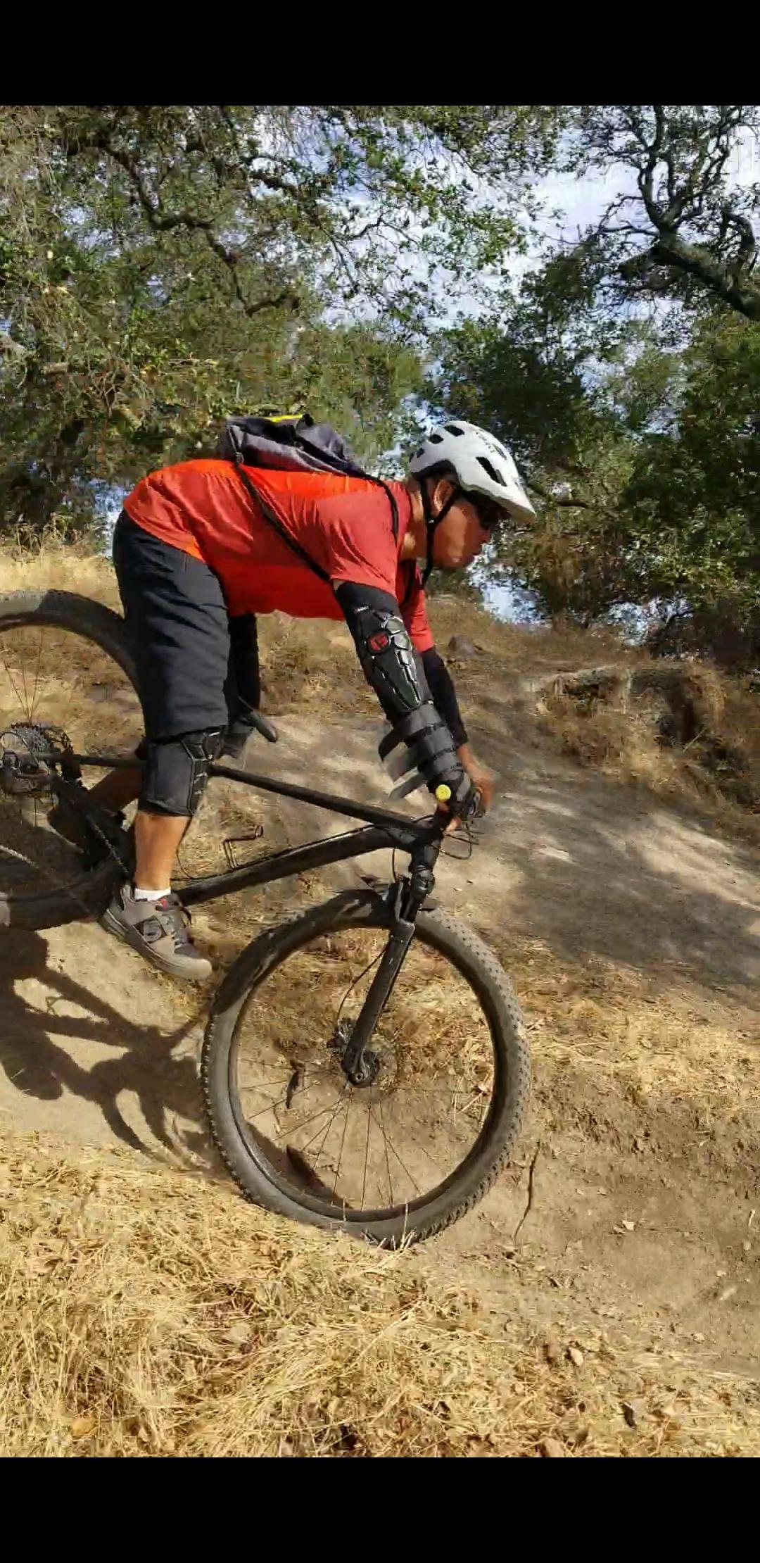 A person in a red shirt and black shorts is riding a mountain bike down a dirt trail. They are wearing a helmet and protective gear on their arms and knees. The surrounding area is dry with sparse grass and trees, and the rider appears focused and in motion, navigating the terrain. Rockville Park mountain bike trail.