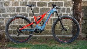 Specialized Specialized Turbo Levo Expert: A mountain bike with a blue and red frame, featuring large tires and a sturdy design, is displayed against a stone wall.