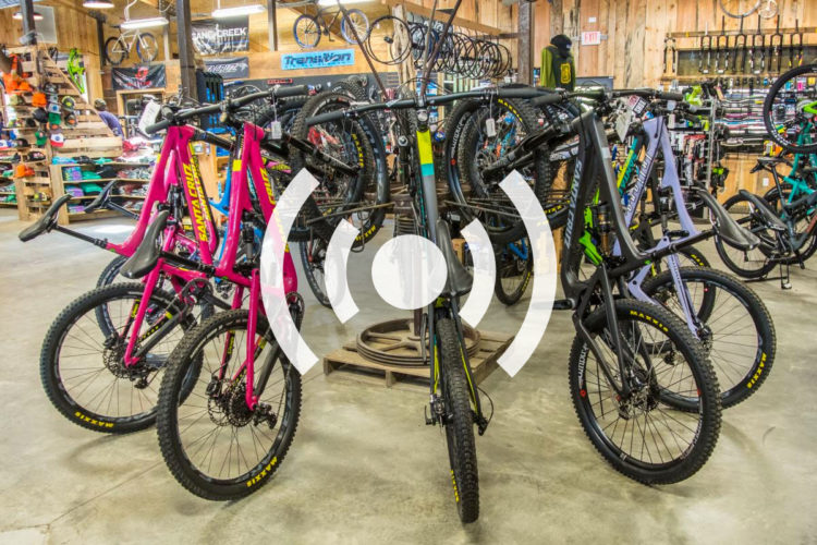 A vibrant display of mountain bikes in a bicycle shop, featuring a variety of colors including pink, green, and black, with accessories and gear visible in the background. The bikes are arranged on a sales floor with wooden accents and hanging bicycle wheels.