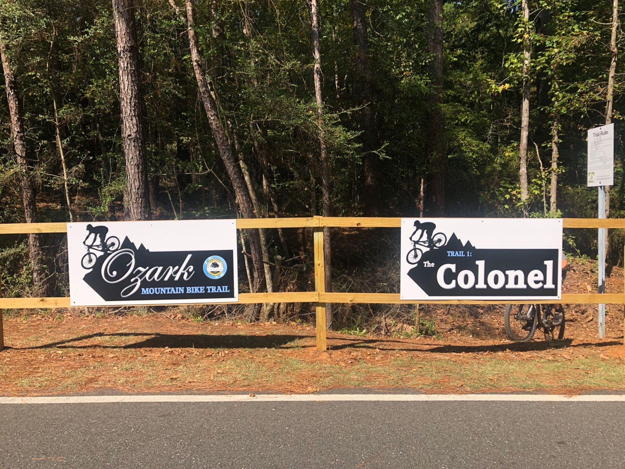 Signage for the Ozark Mountain Bike Trail, featuring two signs: one for the main trail and another for Trail 1, named "The Colonel." The background includes dense trees and a bike leaning against the wooden fence. Ed Lisenby Lake mountain bike trail.