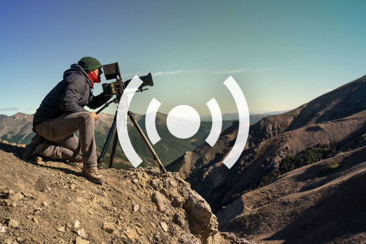 A photographer kneels on rocky terrain, using a video camera mounted on a tripod to capture the panoramic view of a mountainous landscape under a clear blue sky.