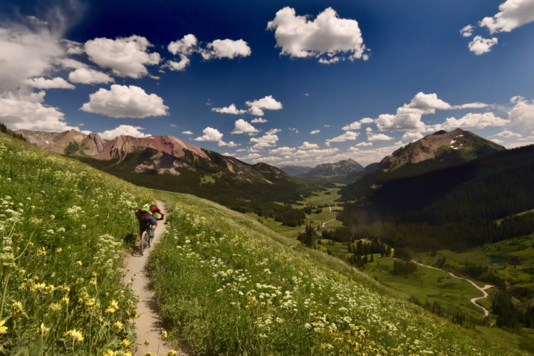 A scenic mountain landscape featuring a winding dirt trail surrounded by lush green grass and wildflowers. A cyclist rides along the trail, with expansive valleys and mountains in the background under a bright blue sky filled with fluffy white clouds.