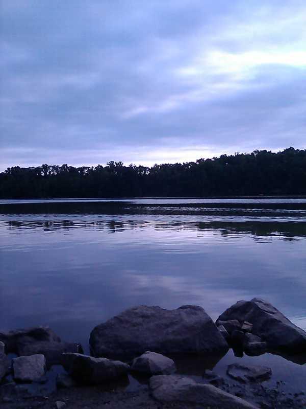 A tranquil landscape featuring a calm river reflecting the overcast sky, bordered by trees in the background, and large rocks in the foreground. Marsh Creek Park mountain bike trail.