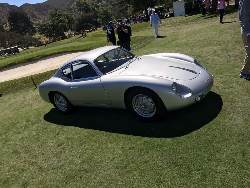 A classic silver sports car is parked on a grassy area, surrounded by people enjoying a sunny day. In the background, a golf course and trees are visible, while onlookers gather, some taking photos. The car has a sleek design with rounded features and a shiny finish.