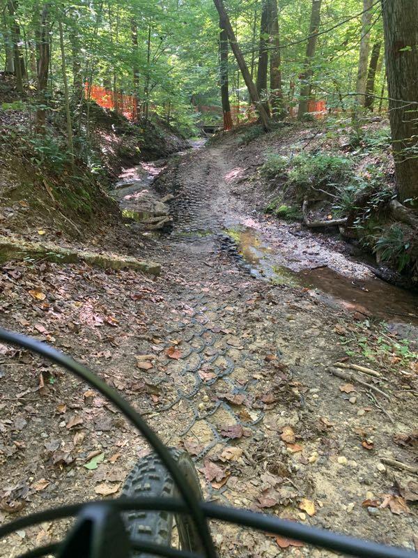 A bike trail winding through a wooded area, featuring a rocky path and patches of shallow water. The surroundings are lush with green foliage, and some sections of the trail are marked with orange fencing. The view is from the perspective of a cyclist, showing part of the bike in the foreground. Rosaryville State Park mountain bike trail.