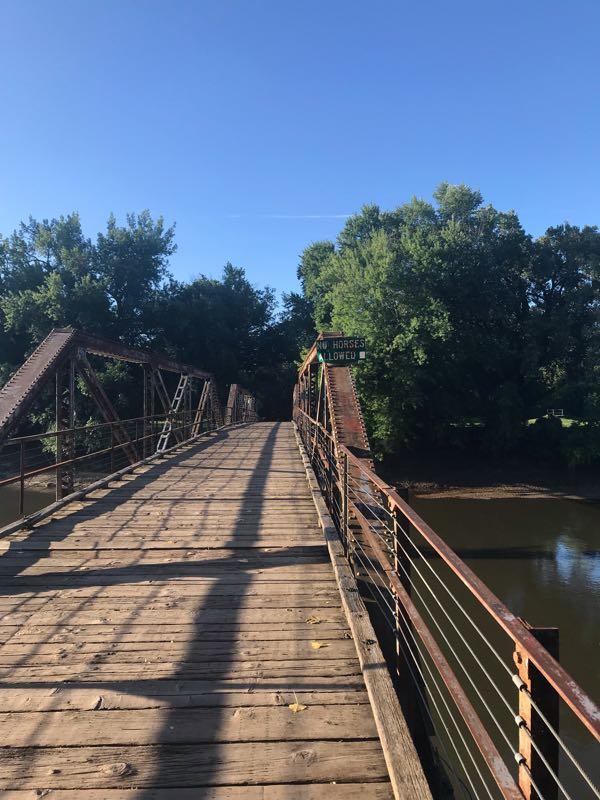 A wooden bridge with a metal frame spans over a river, surrounded by lush greenery. The bridge is mostly empty, with a sign indicating that horses are allowed. The scene is bright, showcasing a clear blue sky and sunlight casting shadows on the wooden planks. The Center Trails mountain bike trail.