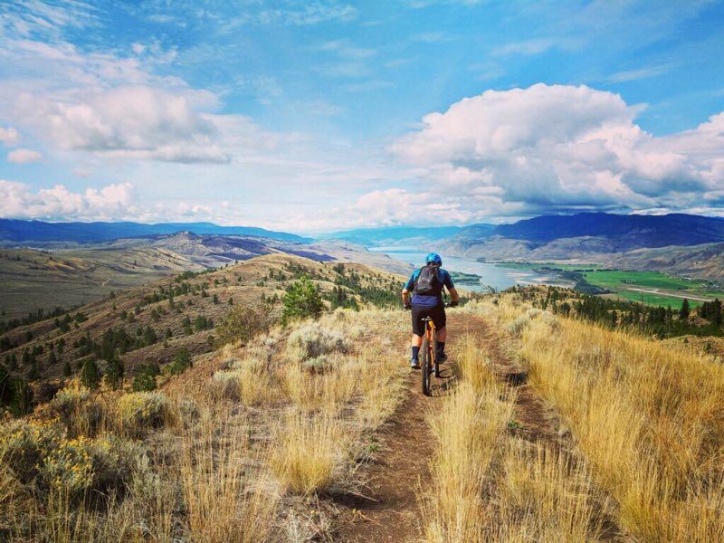 A mountain biker riding along a dirt trail on a hillside, surrounded by tall grass and sparse trees. The view reveals a vast landscape with rolling mountains and a river in the valley below, under a blue sky scattered with clouds. Kenna Cartwright Park mountain bike trail.