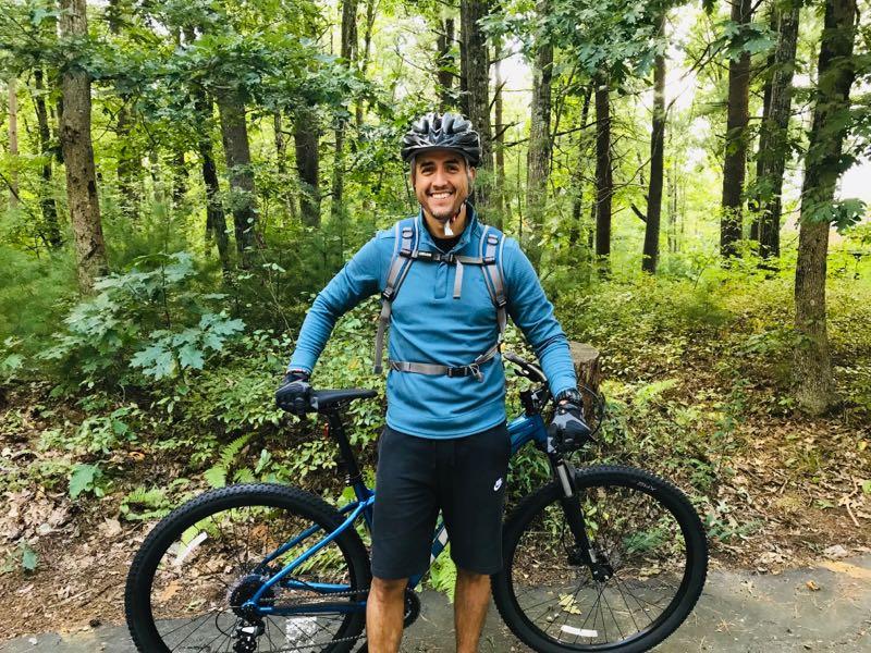 A smiling young man stands next to his blue mountain bike, wearing a helmet and a blue athletic jacket, in a lush forest setting. He has a backpack and gloves, with green trees and foliage surrounding him, suggesting he is ready for a bike ride in nature. Harold Parker State Forest mountain bike trail.