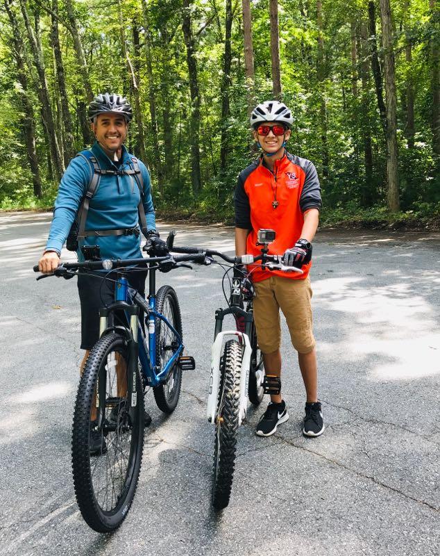 Two cyclists stand next to their bikes on a paved path surrounded by trees. One cyclist, wearing a blue jacket and helmet, smiles while holding a mountain bike. The other cyclist, dressed in a red and black shirt, also wears a helmet and stands next to a white bike, equipped with a camera on the handlebars. Both appear to be enjoying a sunny day outdoors. Harold Parker State Forest mountain bike trail.