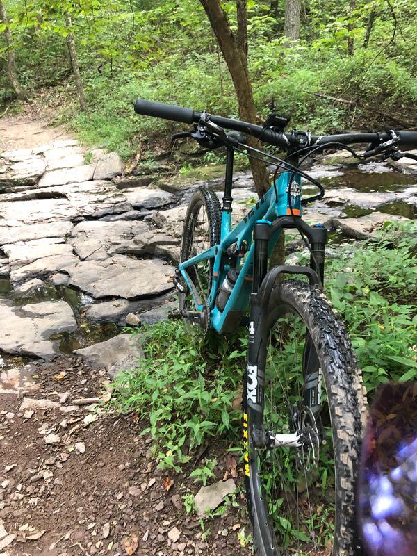 A blue mountain bike parked near a rocky creek in a lush green forest. The bike is positioned on a dirt path, with large stones visible in the water. Surrounding vegetation includes trees and undergrowth, indicating a natural outdoor setting. Nockamixon State Park mountain bike trail.