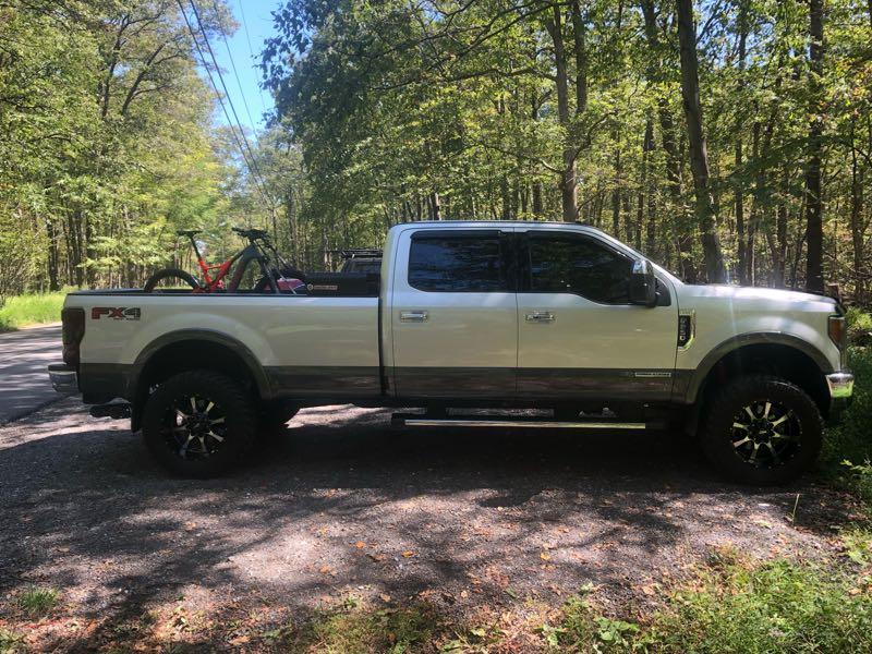 A large white pickup truck parked on a gravel road surrounded by trees. The truck has a bike rack in the bed with a mountain bike secured, and the sun is shining through the foliage, creating dappled light on the ground. Gambrill State Park mountain bike trail.