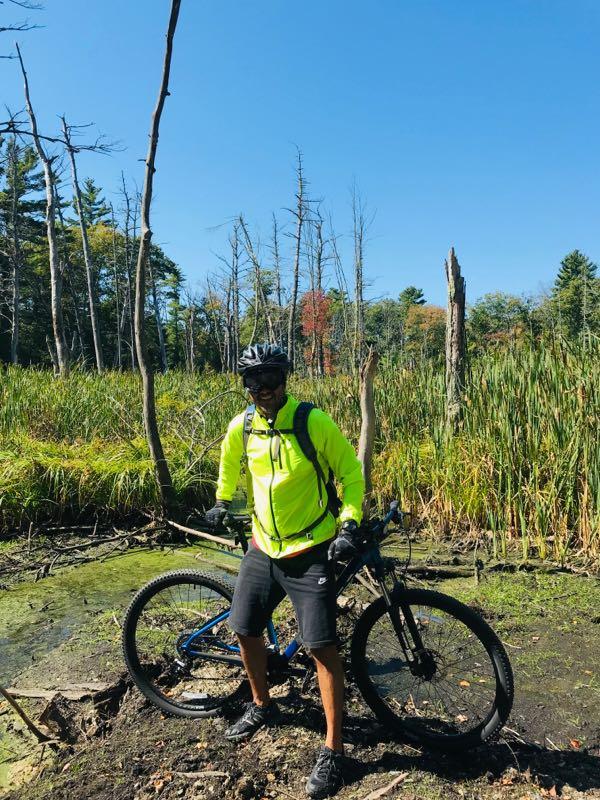 A person wearing a bright yellow cycling jacket and a helmet stands next to a mountain bike in a marshy area. The background features tall grass, small trees, and a clear blue sky. The individual appears to be enjoying a day of outdoor biking. Willowdale Forest mountain bike trail.