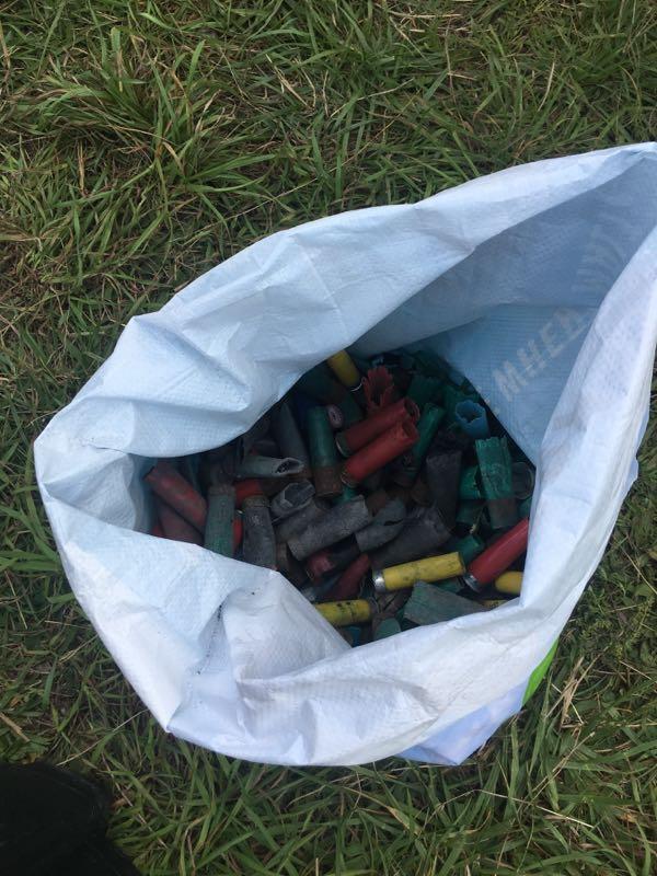 A white bag filled with various colored spent shotgun shells, including red, green, yellow, and black, resting on green grass. Split Rock And Surrounding Area mountain bike trail.