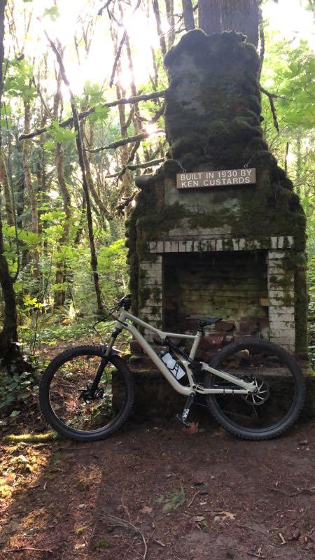 A mountain bike leaning against an old moss-covered stone chimney in a lush green forest. The chimney displays a sign that reads "BUILT IN 1930 BY KEN CUSTARDS." Sunlight filters through the trees, illuminating the scene. Whipped creek regional park mountain bike trail.