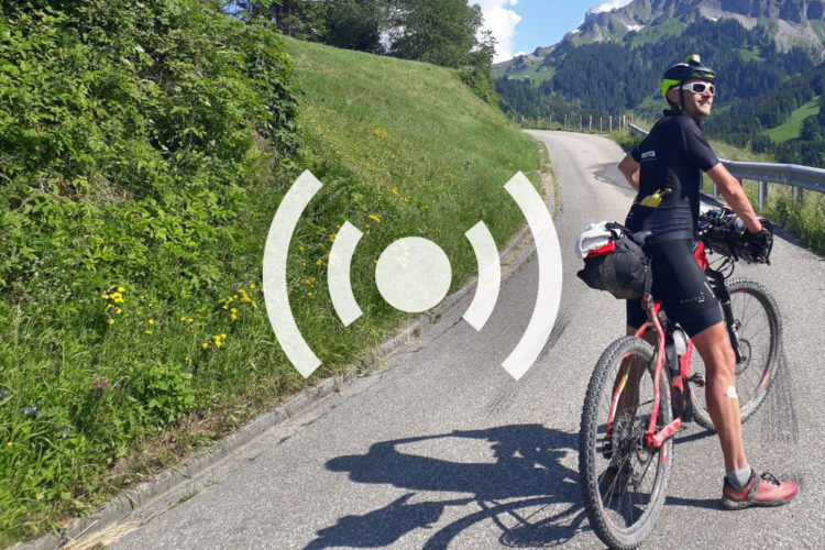 A cyclist pauses on a winding mountain road, surrounded by lush green grass and colorful wildflowers. Dressed in cycling attire, wearing sunglasses and a helmet, they smile while resting on their bike, which is equipped with luggage. Tall trees and majestic mountains form a scenic backdrop under a clear blue sky.