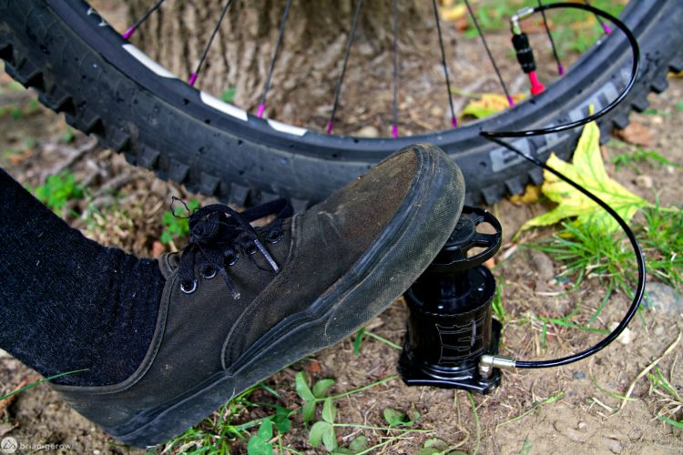 A close-up image of a foot wearing a black sneaker pressing down on a bicycle pump. The bicycle pump is positioned on the ground beside a bicycle tire, with the edge of the tire partially visible in the background. Surrounding the pump and tire are patches of grass and dirt, indicating an outdoor setting.