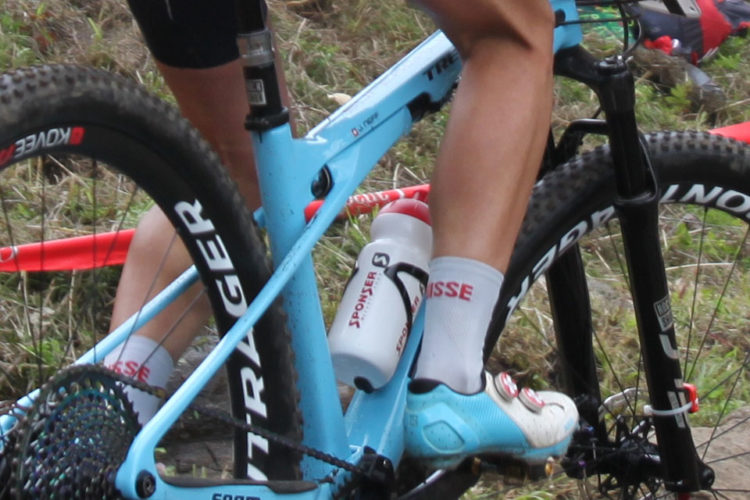 A close-up of a cyclist's foot in a shoe clipped into a bike pedal, accompanied by a light blue bicycle with prominent branding. A water bottle labeled "SPONSER" is visible near the bike frame, with foliage in the background.