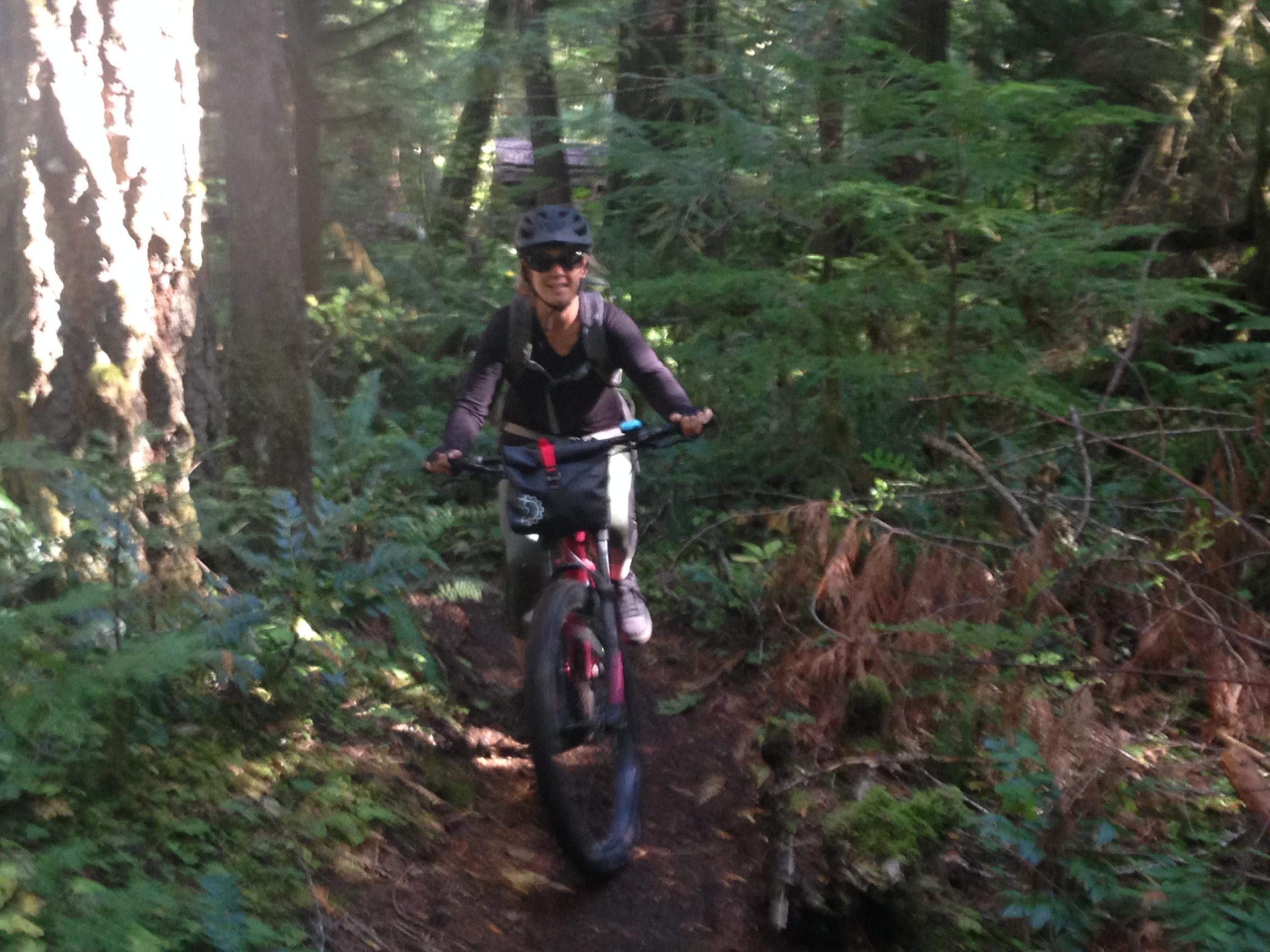 A mountain biker riding through a narrow forest trail surrounded by lush greenery and tall trees, wearing a helmet and sunglasses. The biker is smiling, showcasing a sense of enjoyment in the outdoor activity. Mckenzie River Trail mountain bike trail.