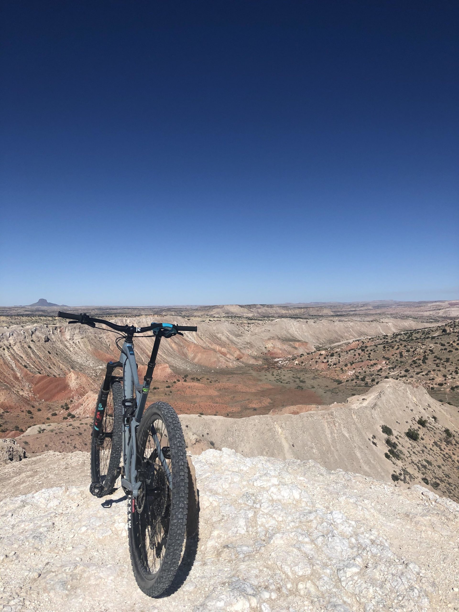 A mountain bike leaning against a rock outcrop, overlooking a vast, arid landscape with layered hills and a clear blue sky. White Ridge Bike Trails mountain bike trail.