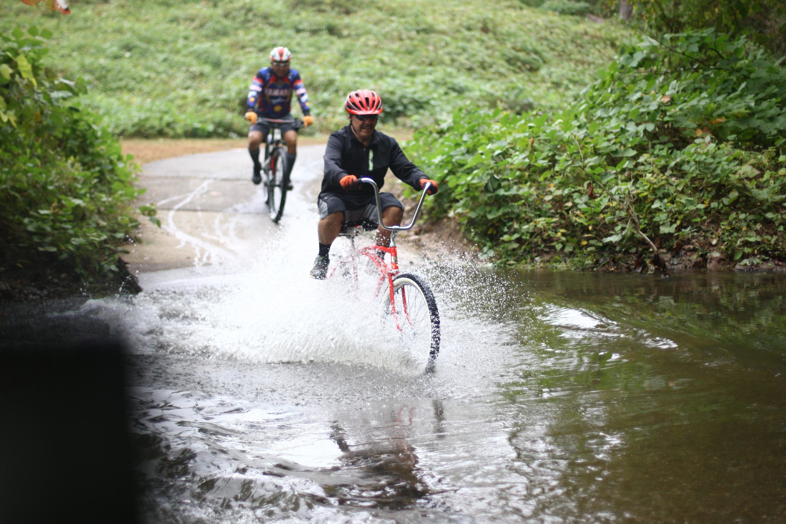 Two cyclists riding on a wet pathway, with one rider splashing through a shallow pool of water. The first cyclist is on a red bike, wearing a helmet and a dark attire. The second cyclist is in the background, on a mountain bike, wearing a patterned jersey and a helmet. Surrounding the path is greenery, creating a natural setting. Salem Lake mountain bike trail.