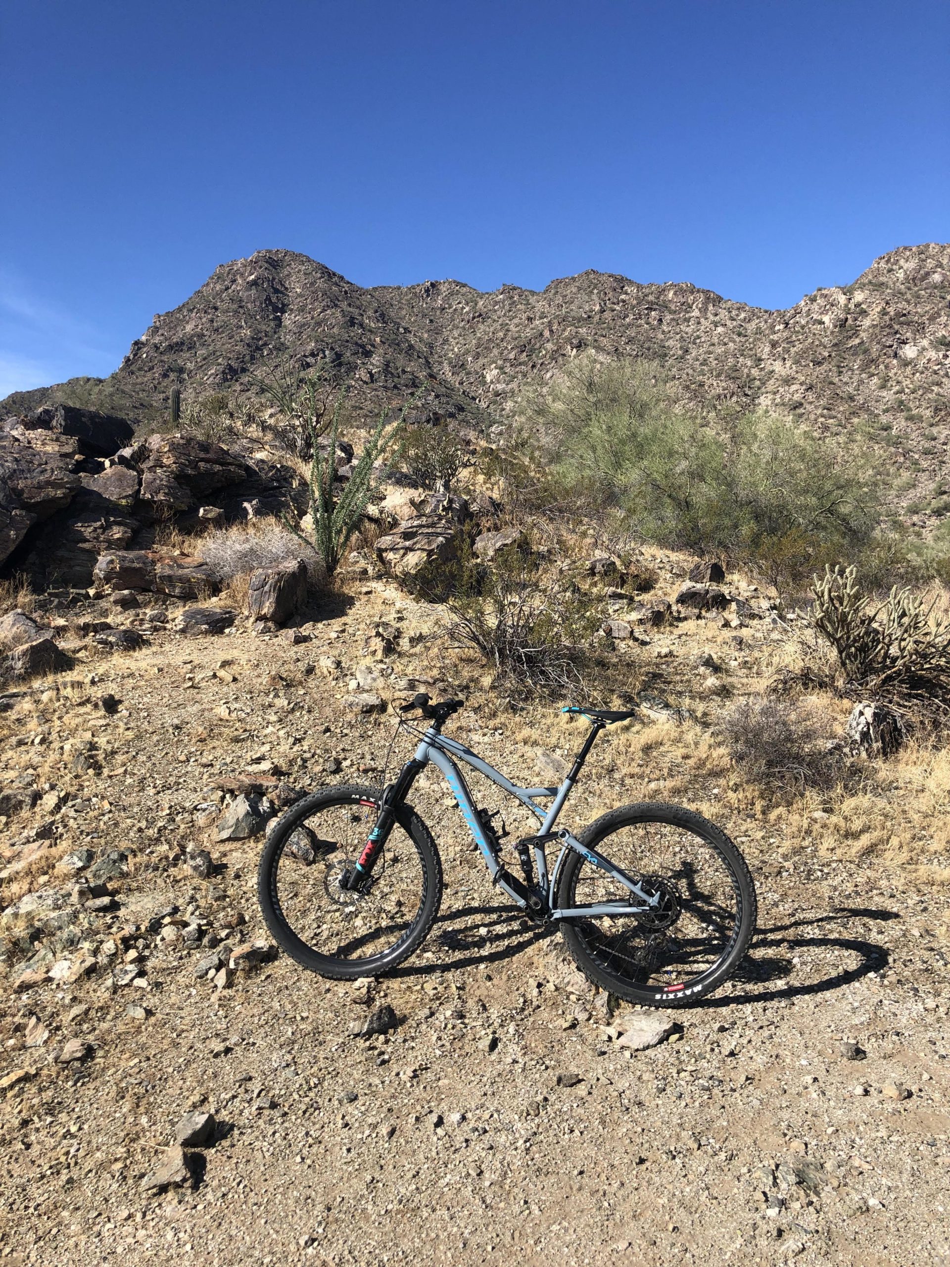 A mountain bike resting on a rocky trail with a backdrop of rugged mountains and a clear blue sky. The terrain features dry vegetation and scattered stones typical of a desert landscape. Estrella Mountain Park mountain bike trail.
